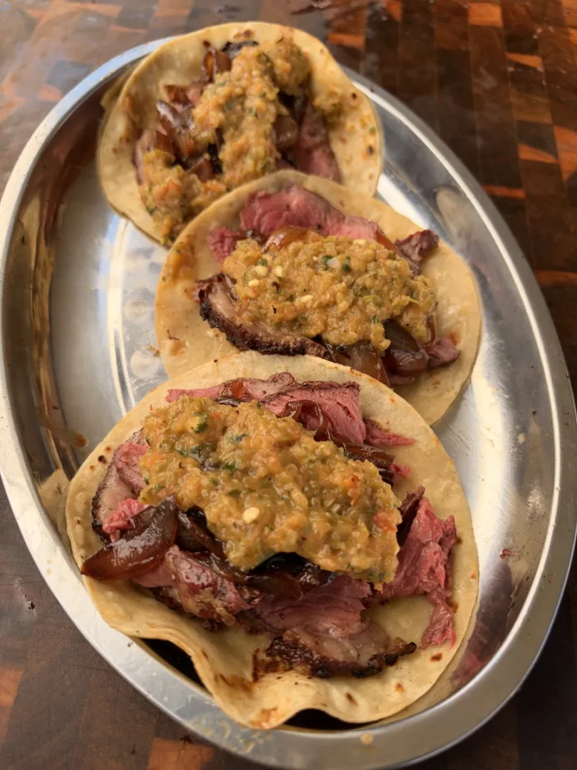 Three corn tortilla tacos filled with thinly sliced rotisserie picanha, sautéed onion, and roasted Hatch chile salsa on a metal platter