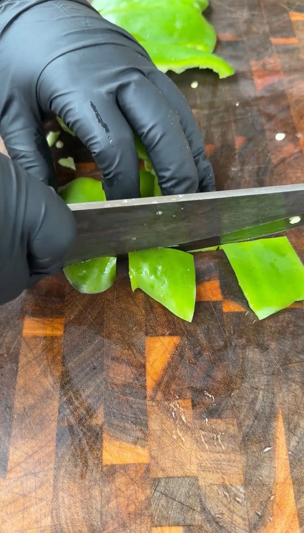 Hands in black gloves roughly chopping green bell pepper into chunky pieces on a wooden cutting board