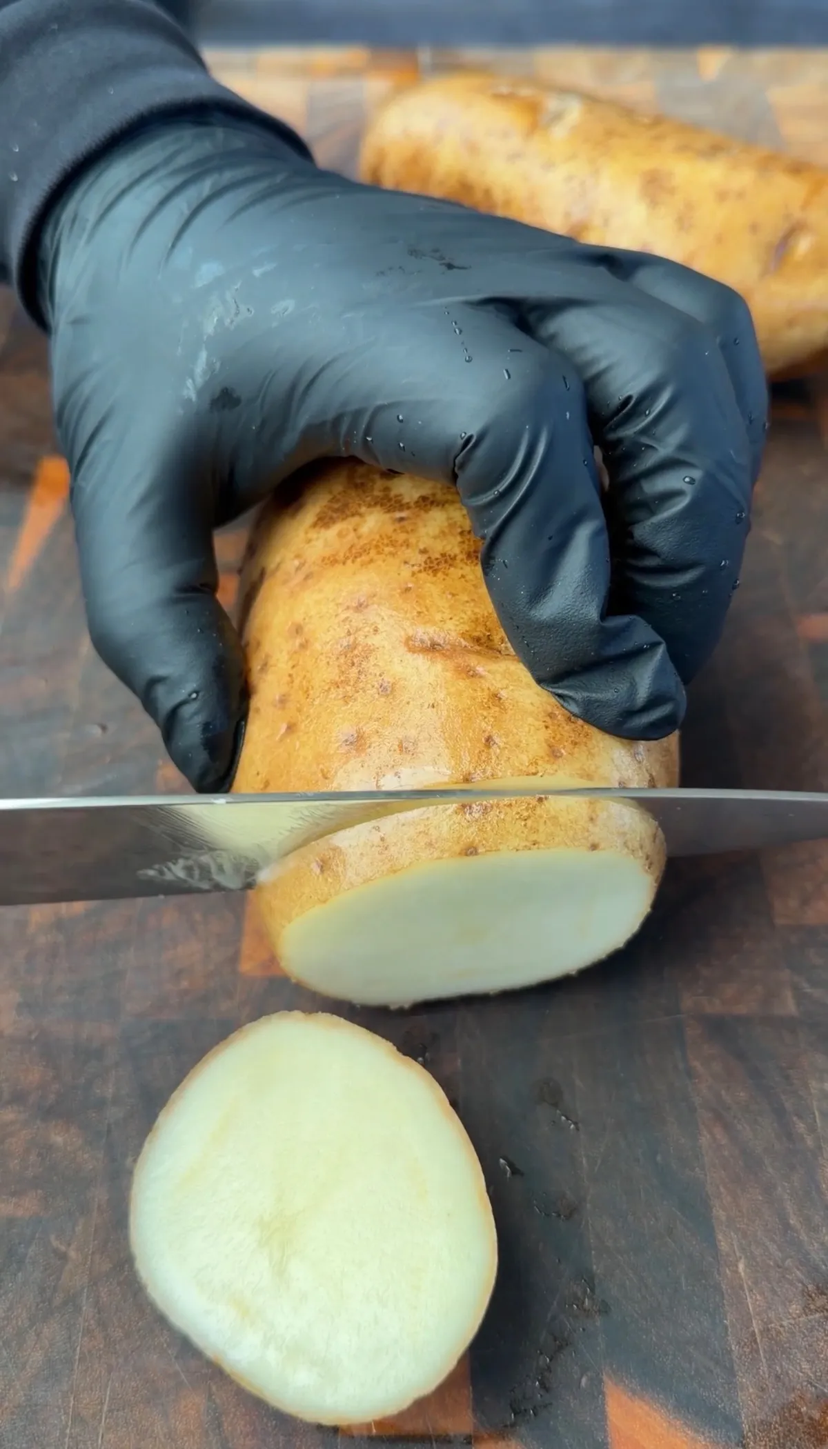 Gloved hands slicing raw russet potatoes into rounds on a wooden cutting board for Grilled Cajun Parmesan Potatoes