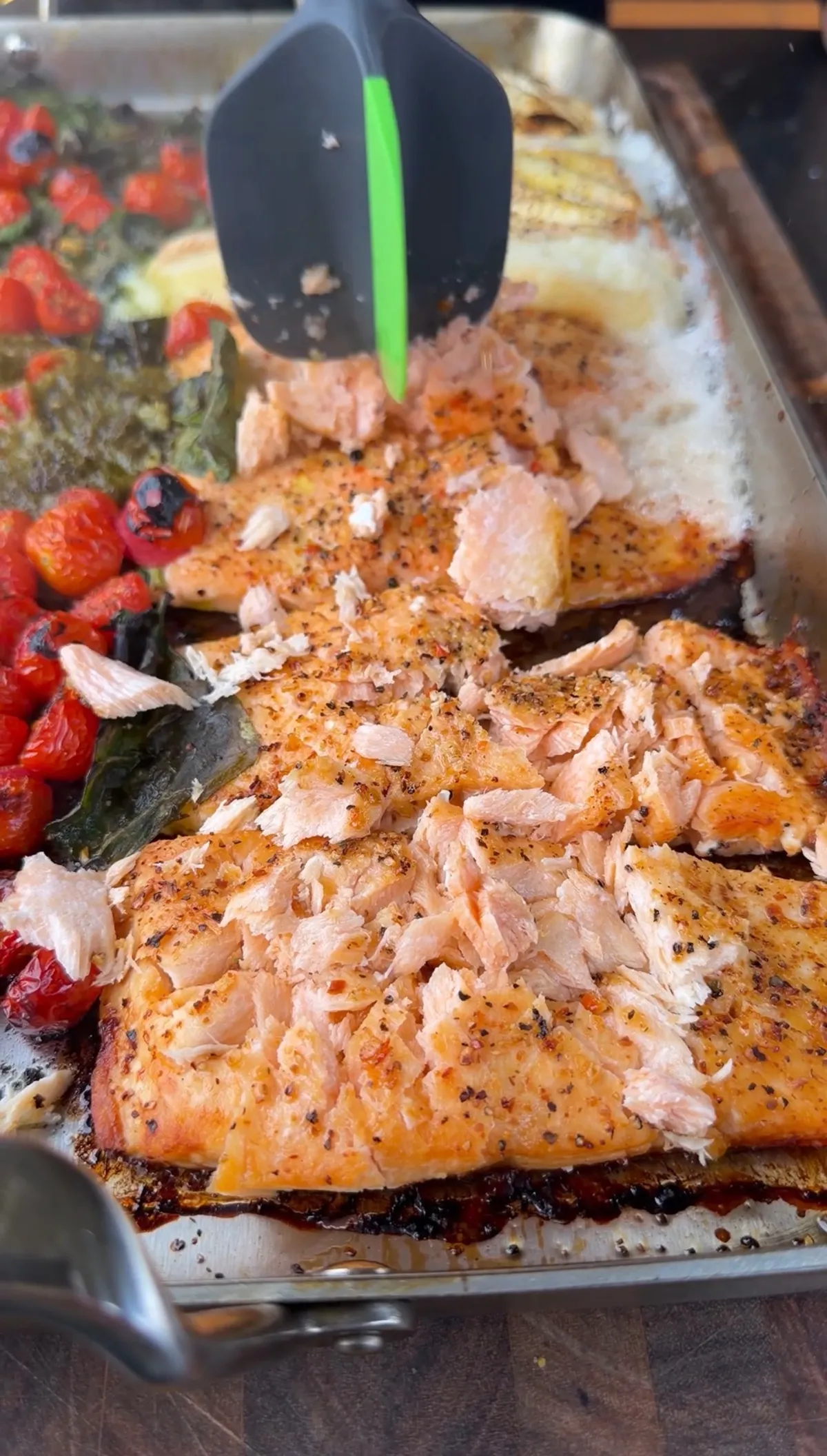 Cooked salmon portions being broken up in a baking pan with roasted cherry tomatoes, wilted spinach, and melted Brie cheese