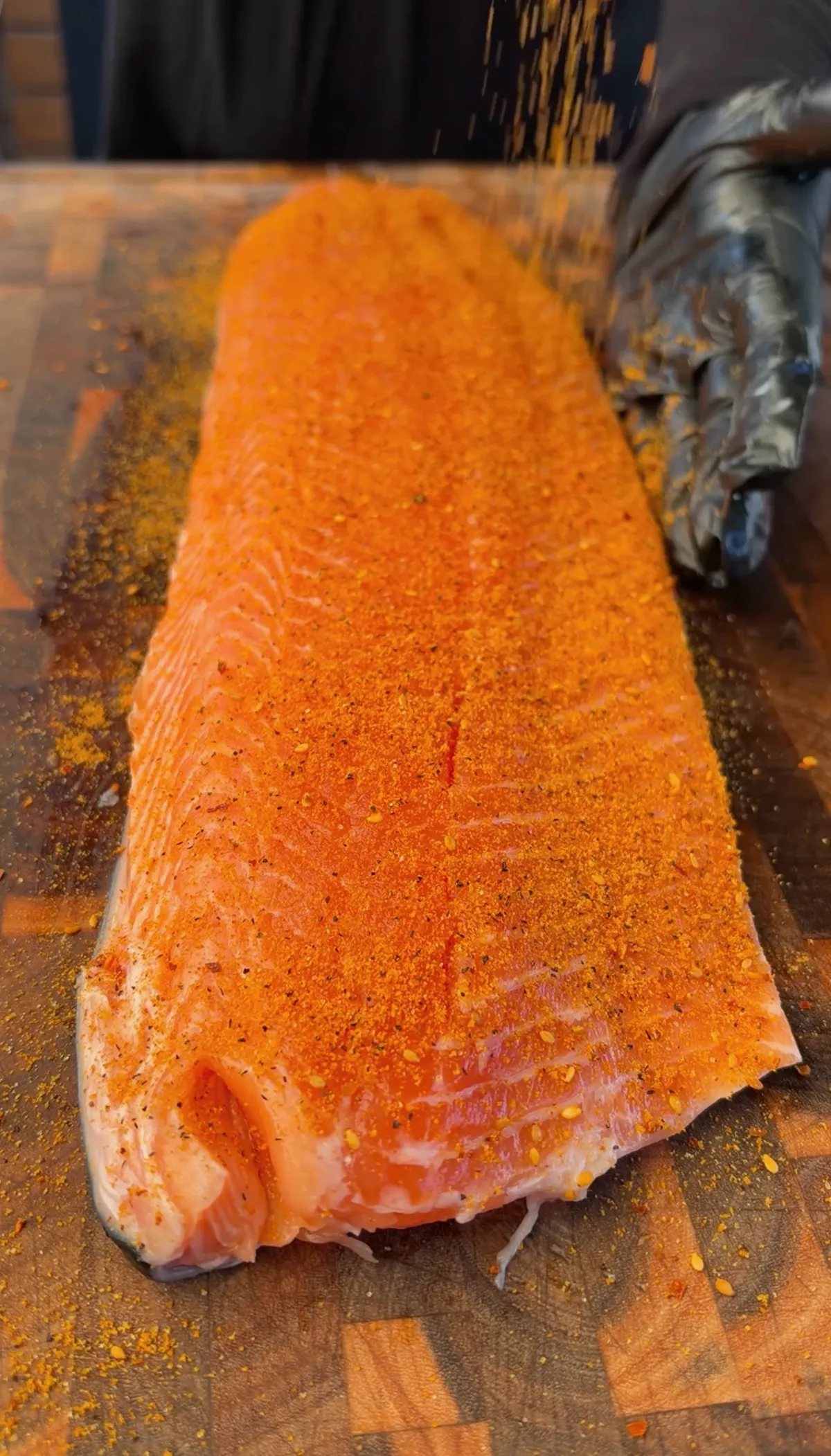 Raw 1-lb salmon fillet being seasoned with seafood seasoning on a wooden cutting board before smoking
