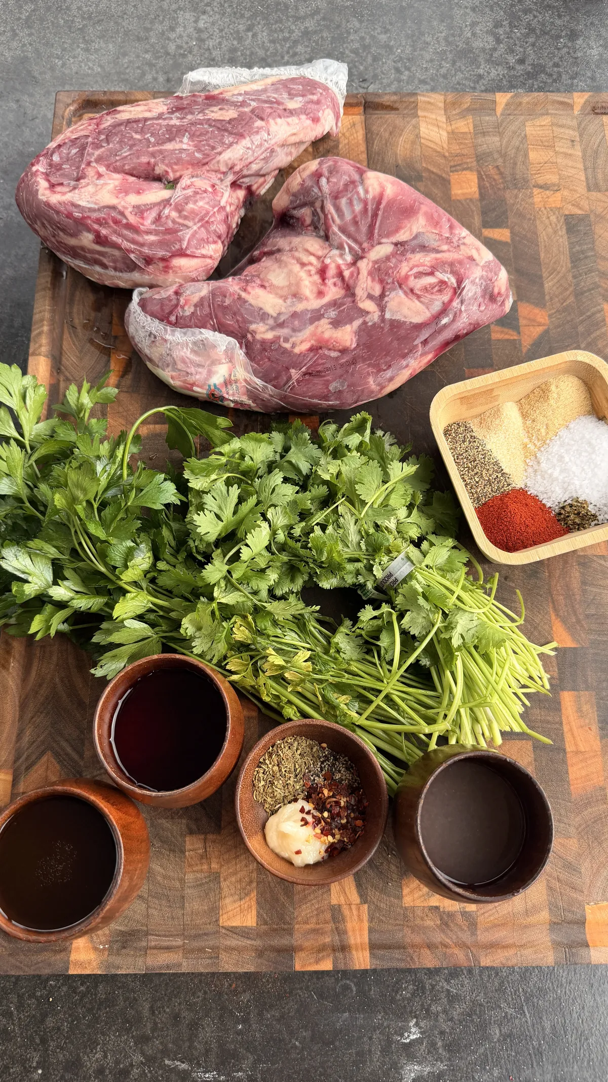 Raw tri-tip with chimichurri ingredients including parsley, cilantro, red wine vinegar, and seasoning mud spices on a wooden board