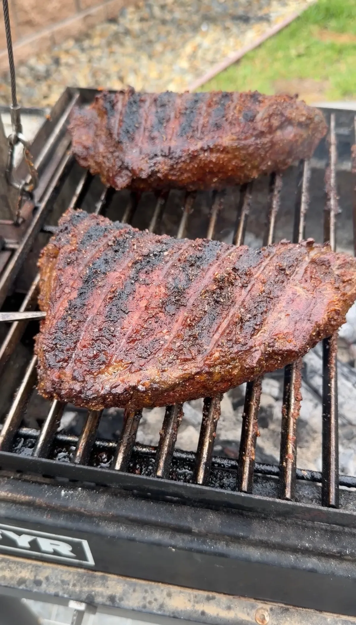 Two seasoned tri-tips with mud rub and rendered fat cap cooking on a Santa Maria grill over charcoal and hickory