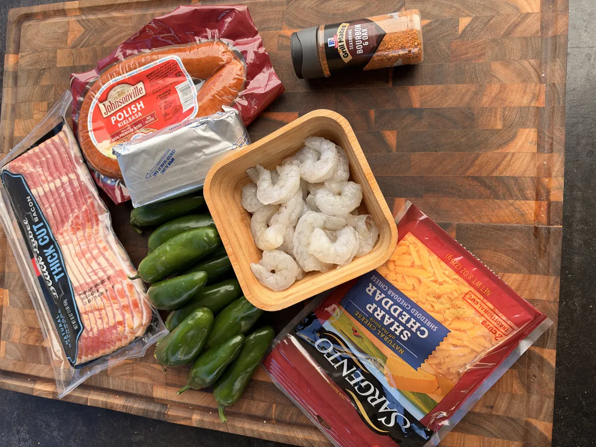 Jalapeño popper ingredients on wooden board: kielbasa, raw shrimp, jalapeños, cream cheese, cheddar, bacon, and BBQ seasoning
