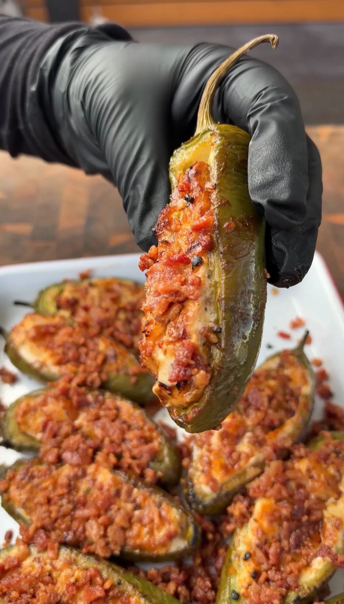 Hand holding smoked jalapeño popper stuffed with sausage, shrimp, cream cheese, cheddar, and sweet BBQ seasoning over a tray of more poppers
