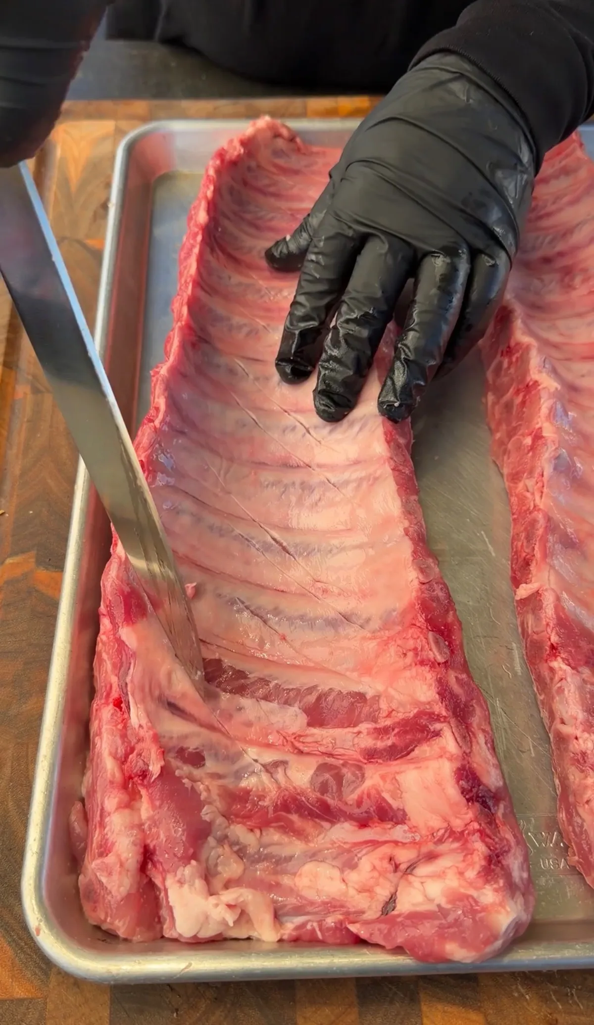 Gloved hands scoring the membrane on raw baby back ribs in a metal tray before seasoning and smoking