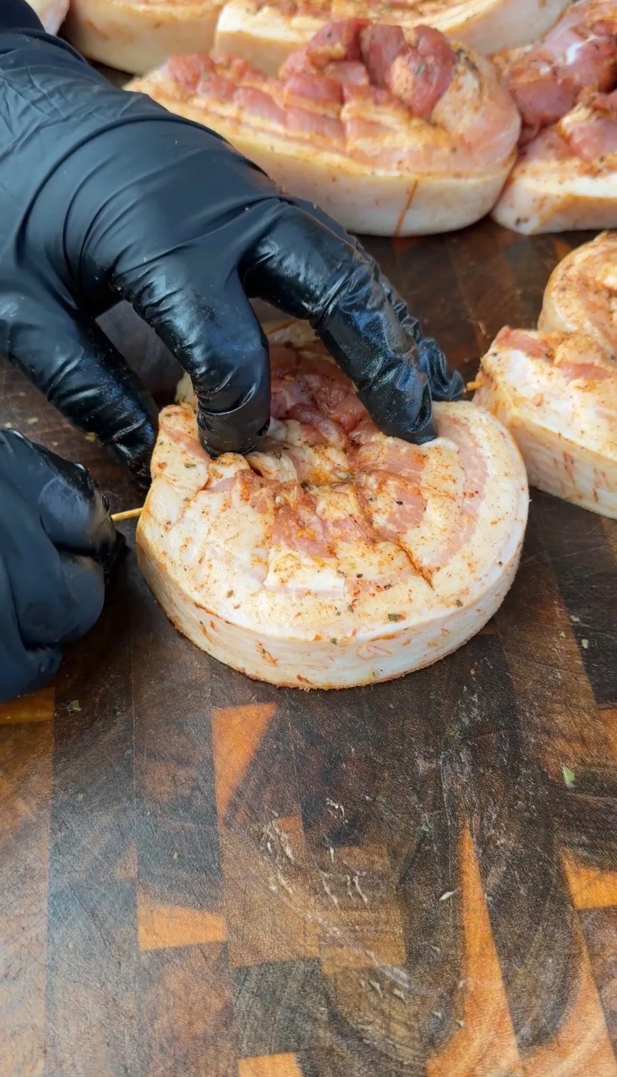 Gloved hands securing a rolled pork belly pinwheel with toothpicks, seasoned with ancho chili seasoning on a cutting board