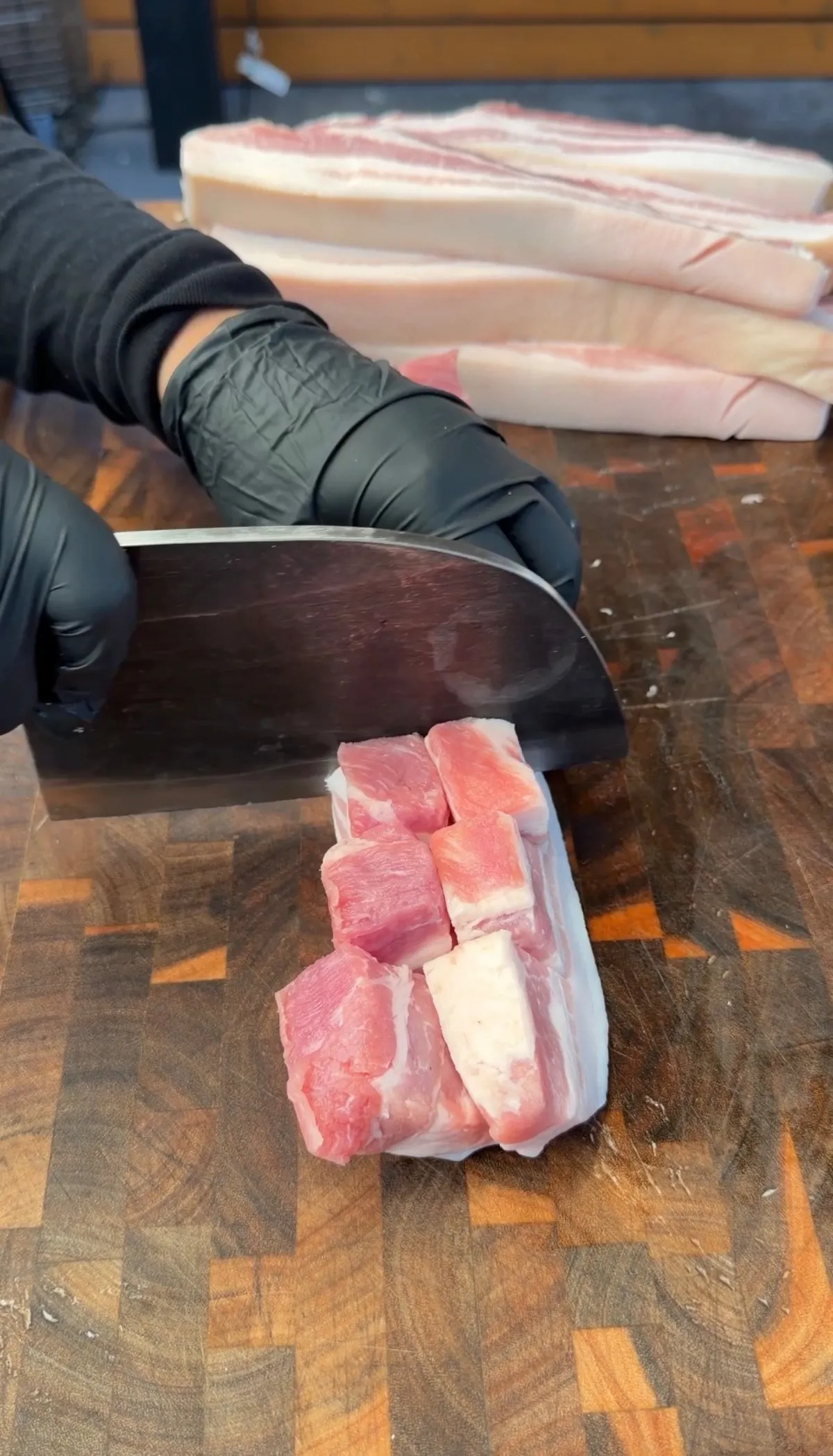 Gloved hands using a sharp knife to score skin-on pork belly strips on an end-grain cutting board for chicharrón pinwheels