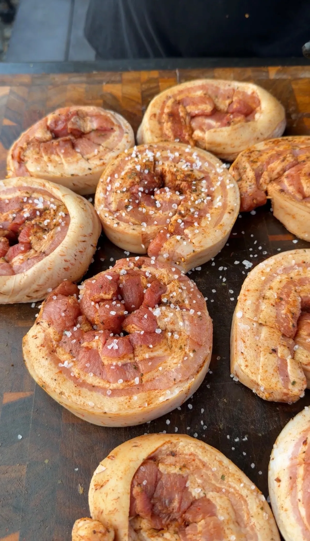 Raw skin-on pork belly pinwheels secured with toothpicks, seasoned with ancho chili seasoning and kosher salt on a wooden cutting board