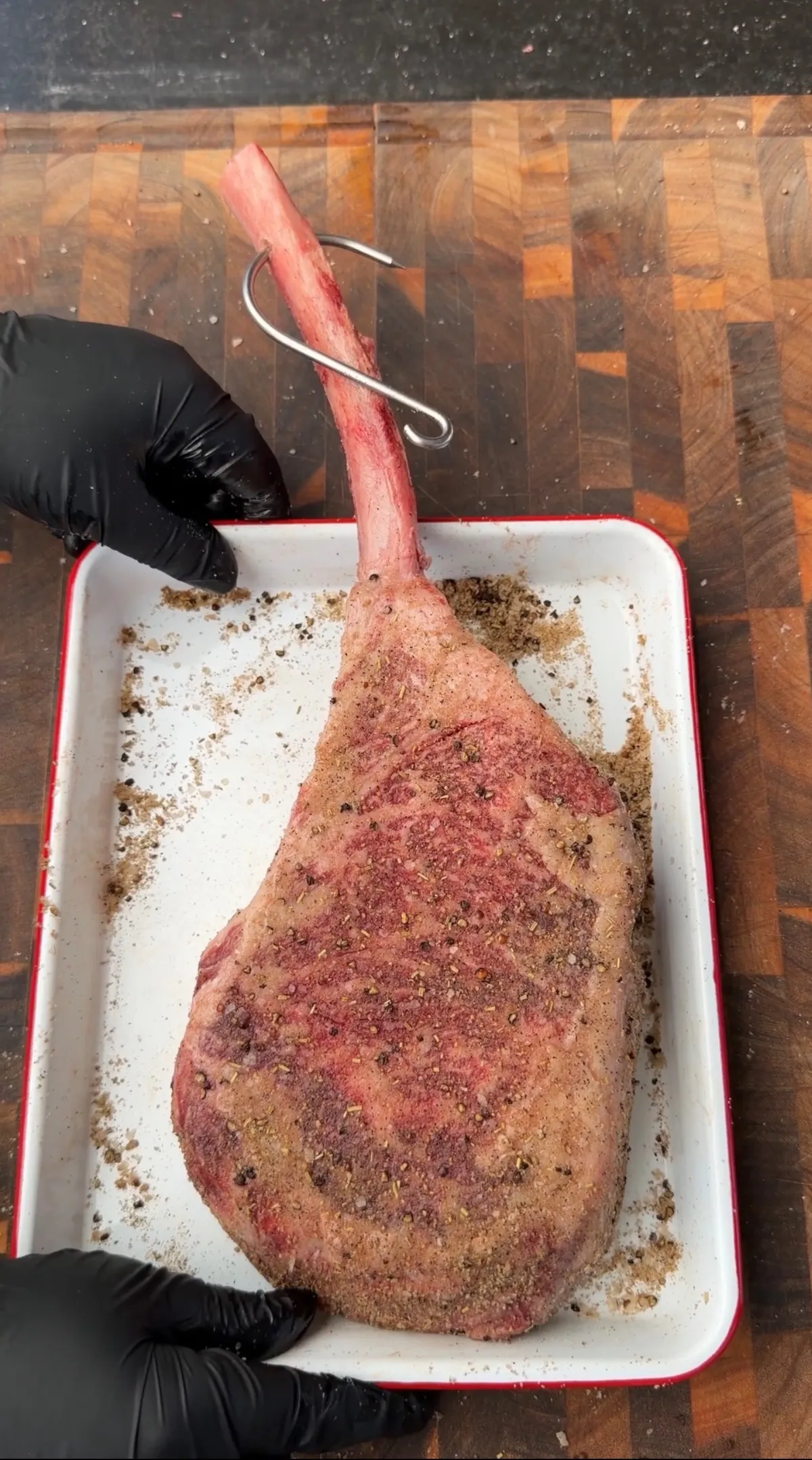 Tomahawk steak seasoned with kosher salt, black pepper, and garlic powder in a white tray with a grill hook through the bone
