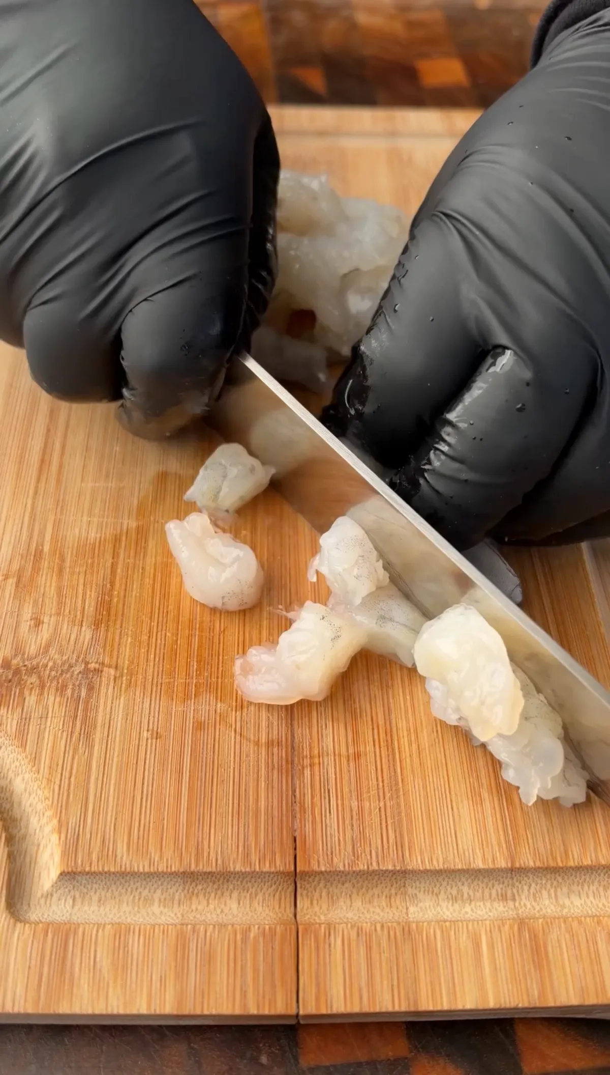 Gloved hands cutting peeled and deveined shrimp into four pieces each on a wooden cutting board