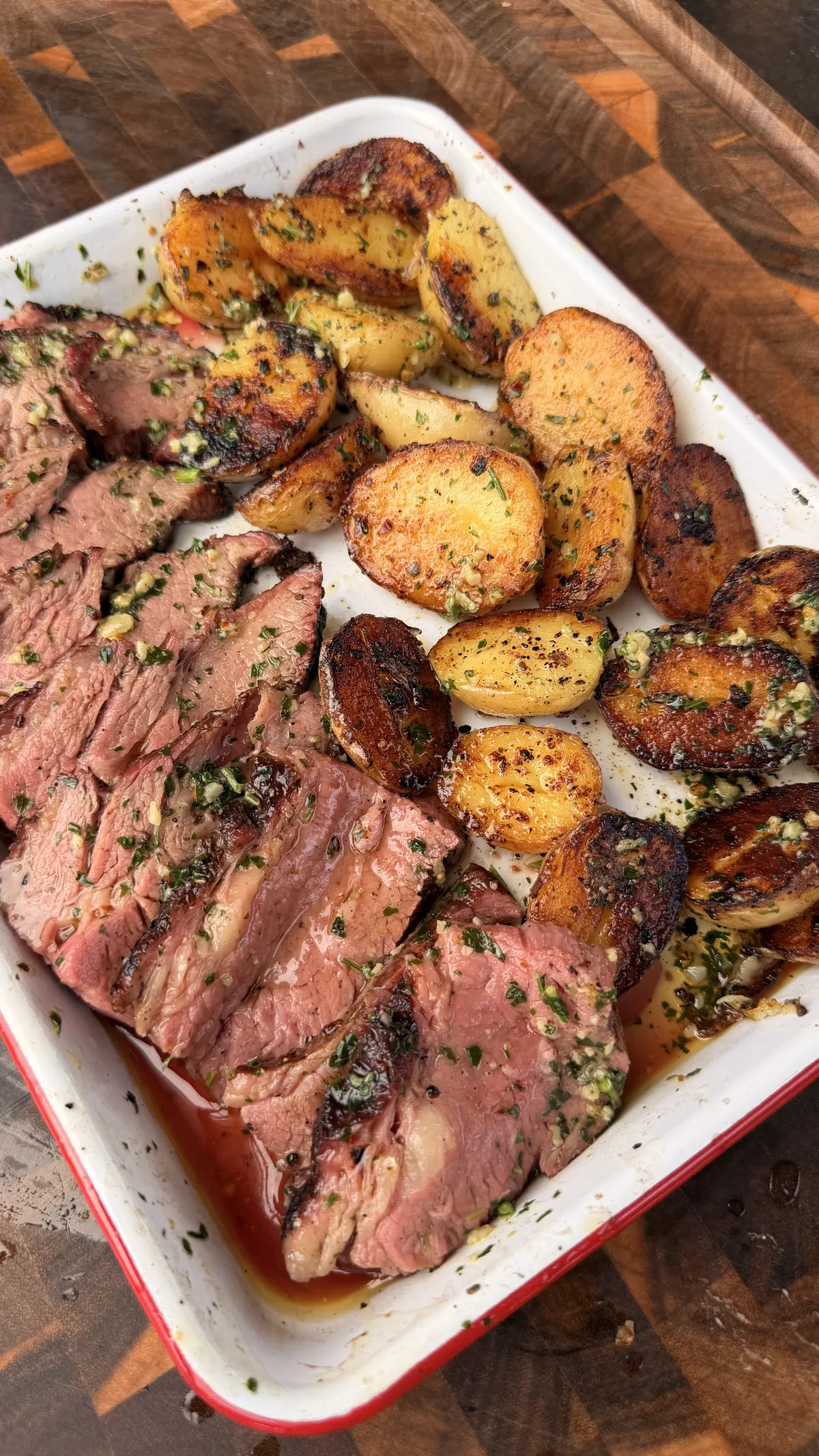 Sliced reverse-seared smoked tri-tip with chimichurri and garlic herb butter alongside crispy roasted lemon pepper potatoes in a white enamel pan