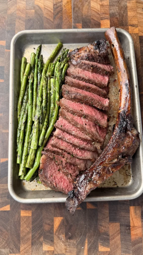 Sliced smoked tomahawk steak with caveman-seared crust served alongside crispy asparagus on a metal tray