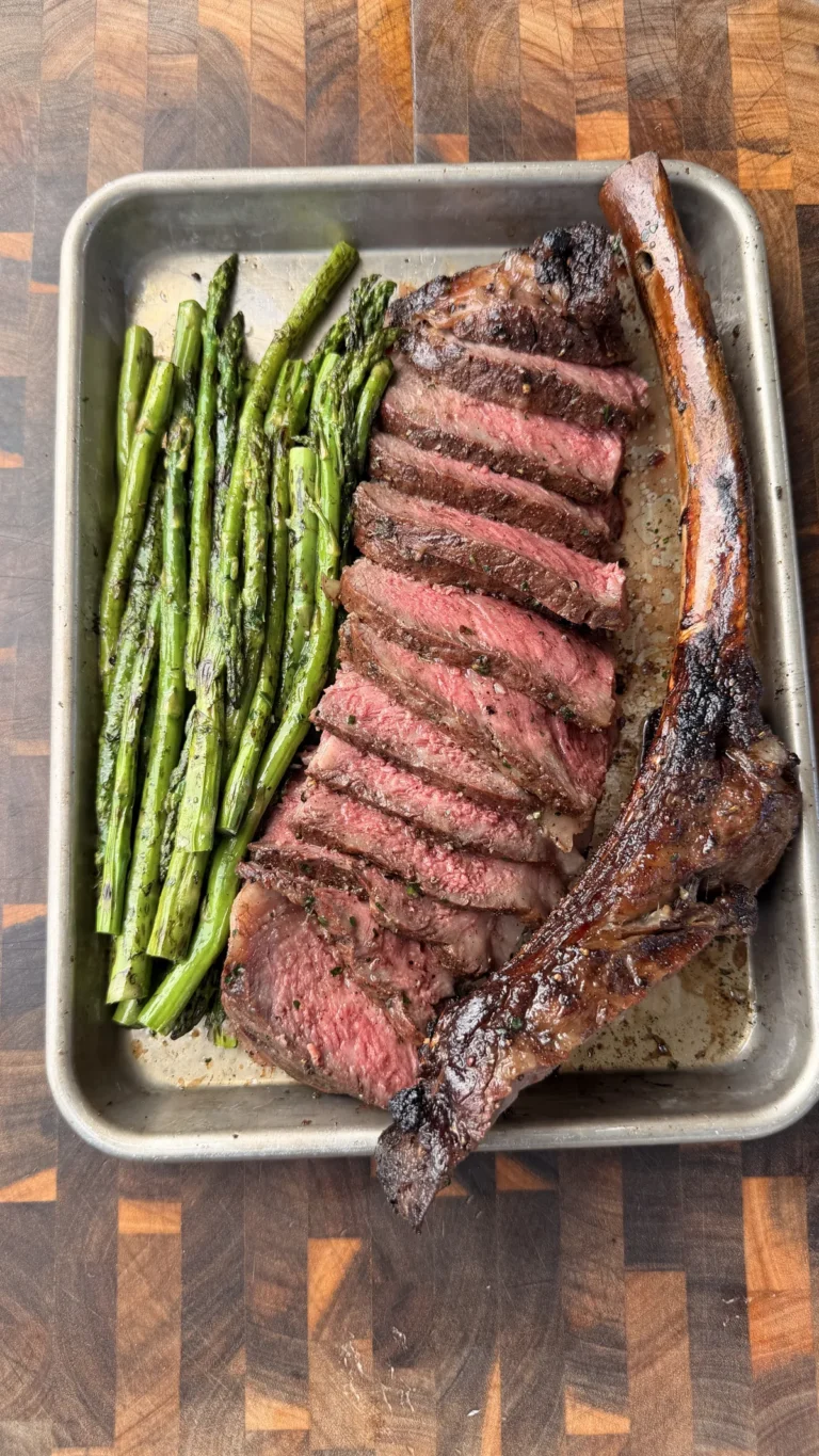 Sliced smoked tomahawk steak with caveman-seared crust served alongside crispy asparagus on a metal tray