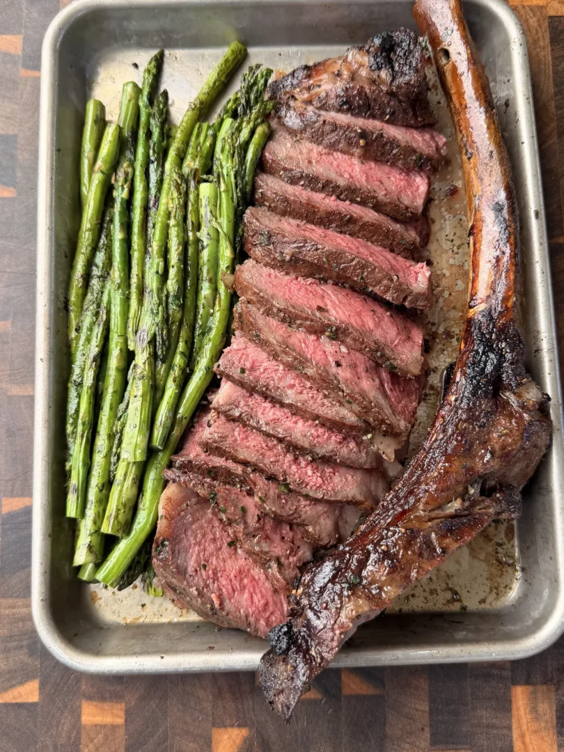 Sliced smoked tomahawk steak with caveman-seared crust served alongside crispy asparagus on a metal tray