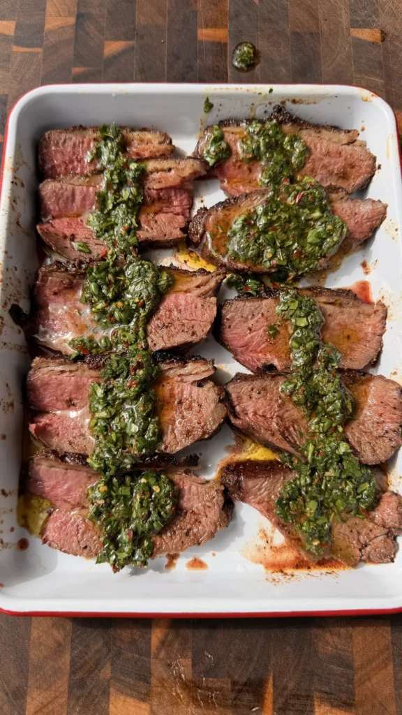 Thinly sliced Santa Maria tri-tip arranged on a white platter, generously topped with fresh parsley and cilantro chimichurri