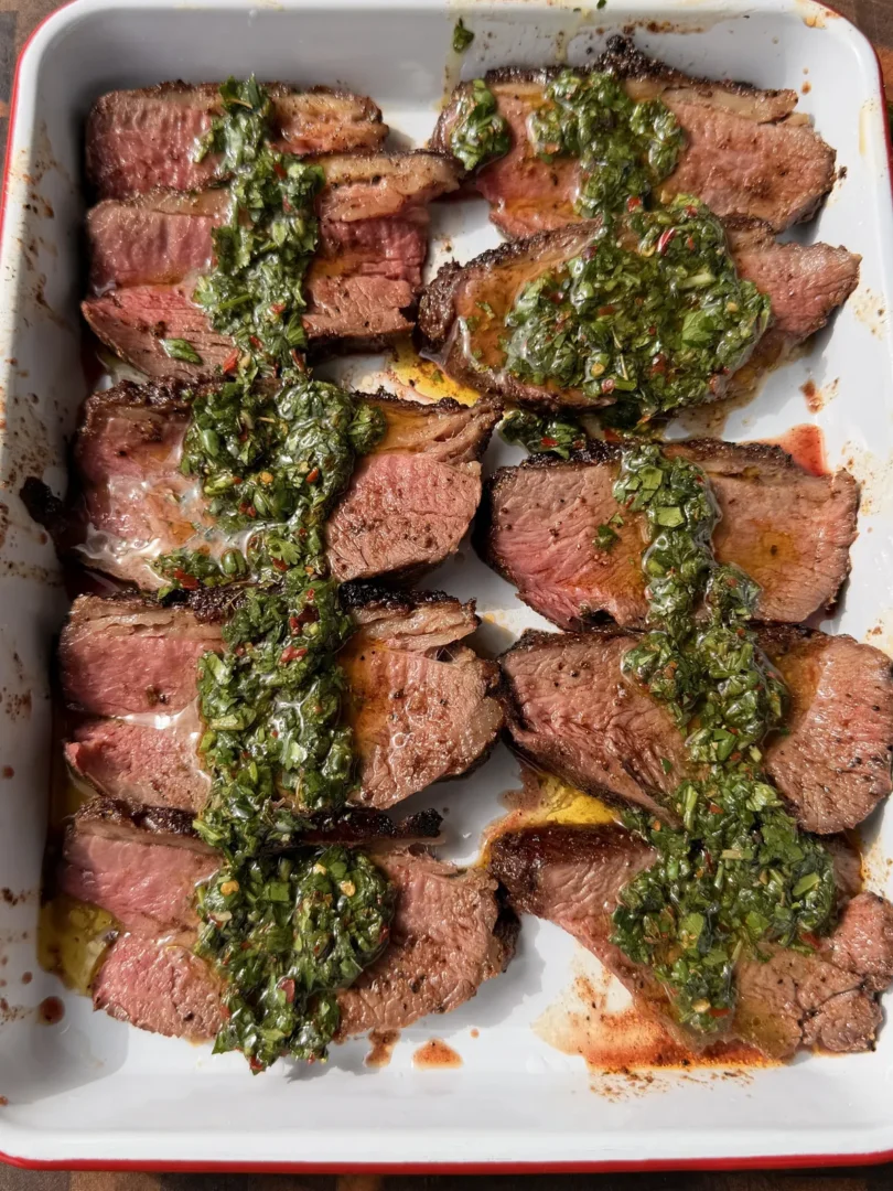 Thinly sliced Santa Maria tri-tip arranged on a white platter, generously topped with fresh parsley and cilantro chimichurri