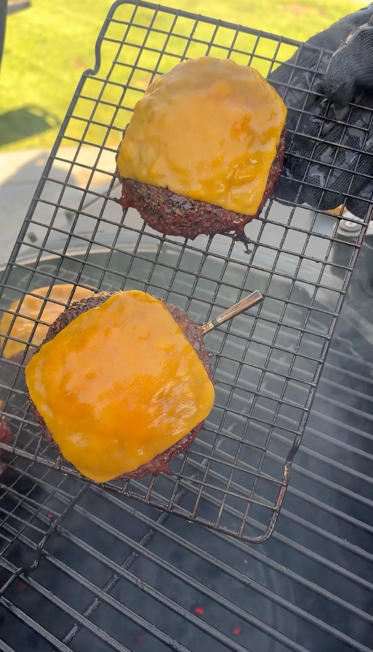 Two smoked beer can burger patties topped with melted cheddar cheese slices resting on a wire rack over a smoker