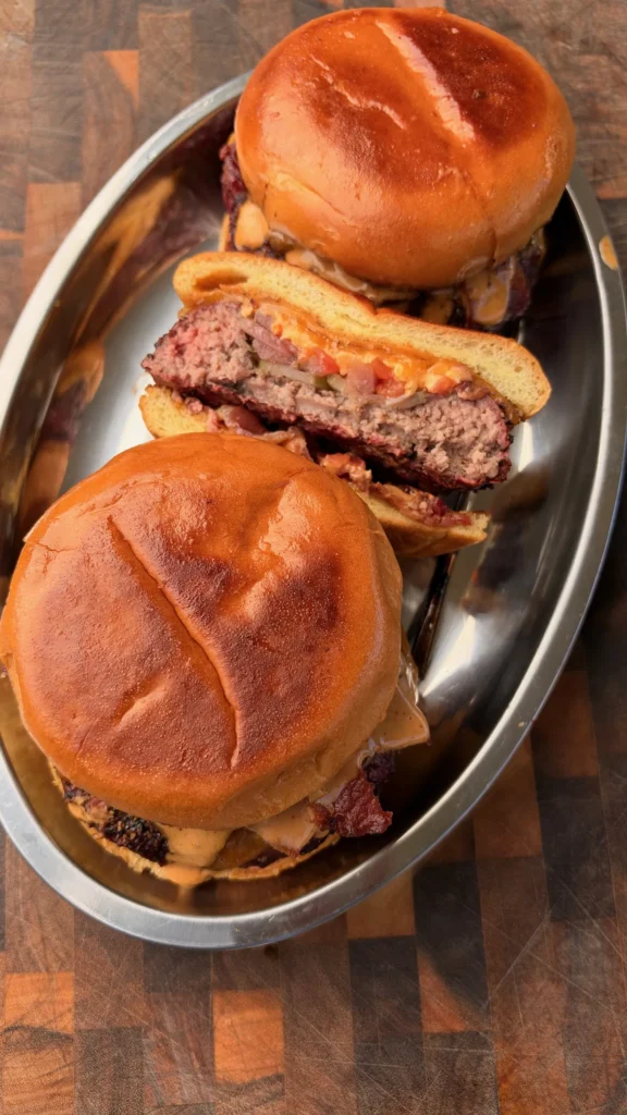 Smoked beer can burgers on brioche buns with chipotle mayo and crispy bacon on a metal tray, one sliced open