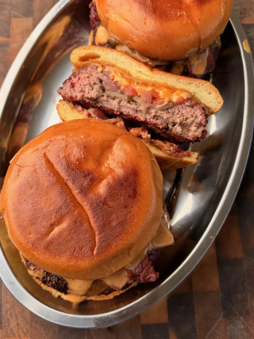 Smoked beer can burgers on brioche buns with chipotle mayo and crispy bacon on a metal tray, one sliced open