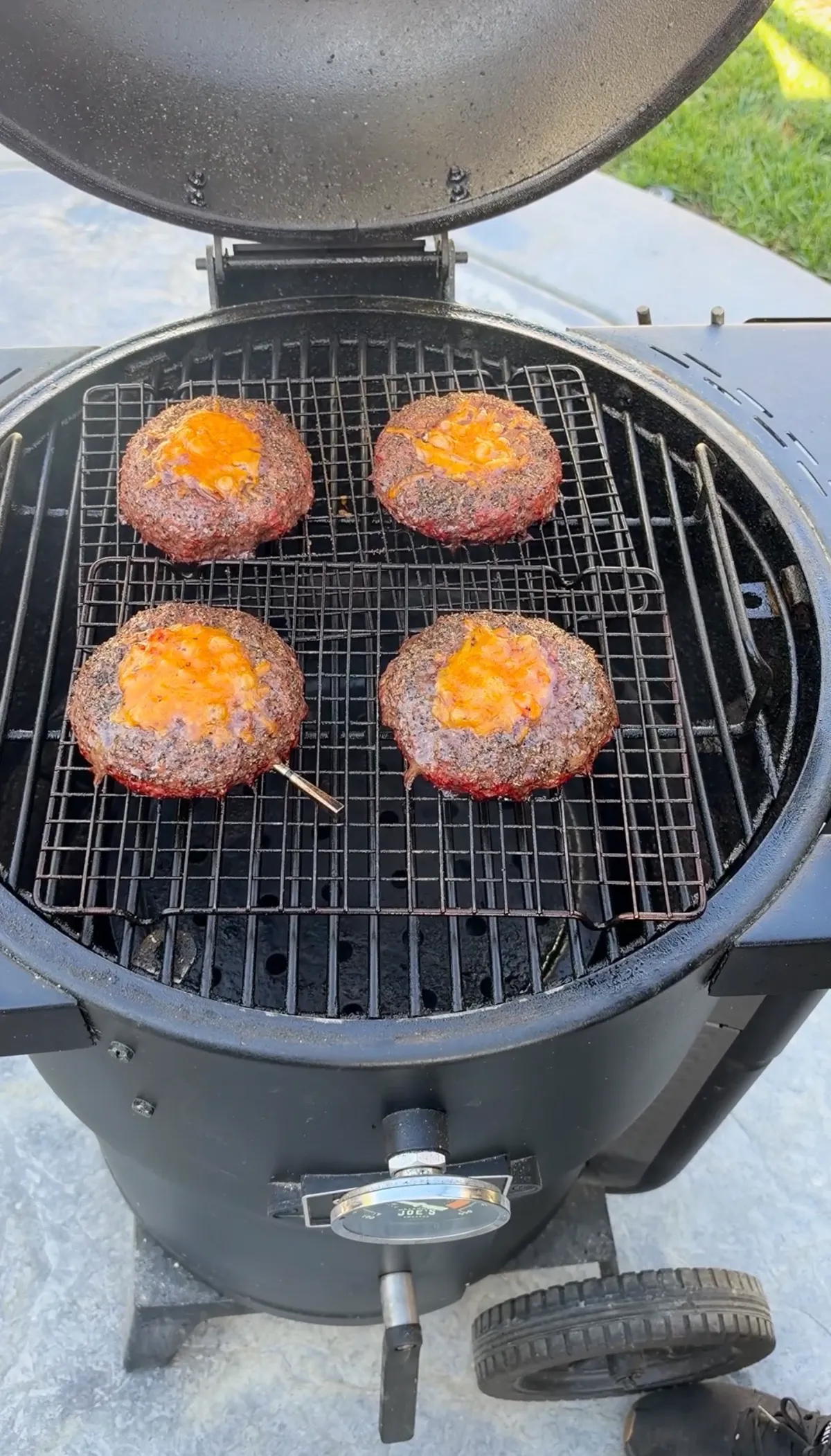 Four stuffed smoked beer can burgers with melted cheddar cheese cooking on a smoker at 250°F