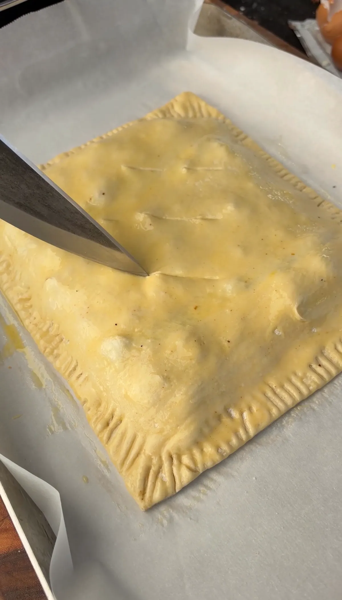 Knife cutting steam slits into egg-washed puff pastry smoked brisket hot pocket on parchment paper
