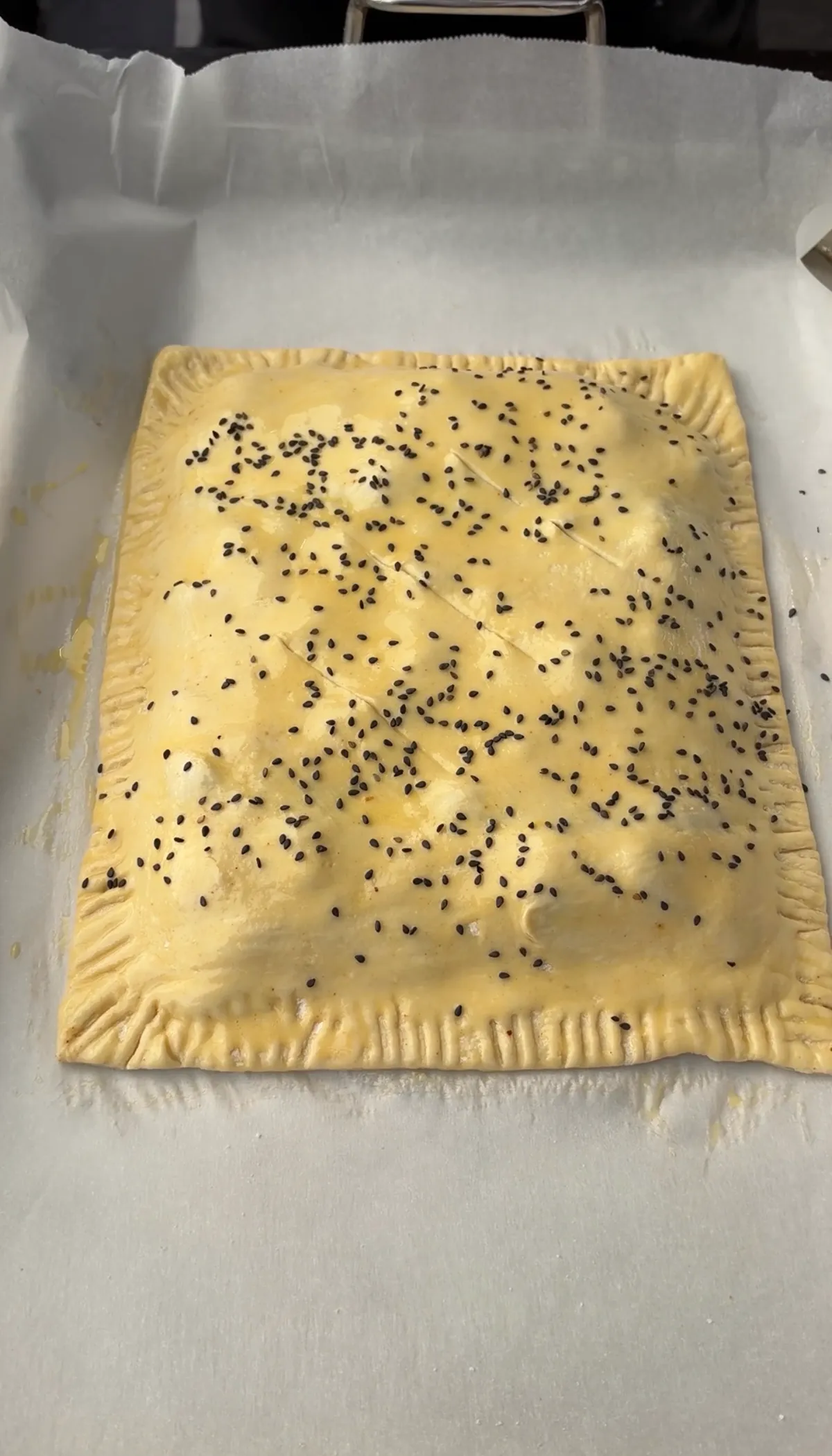 Golden puff pastry brisket hot pocket topped with black sesame seeds, fork-crimped edges, on parchment-lined baking sheet