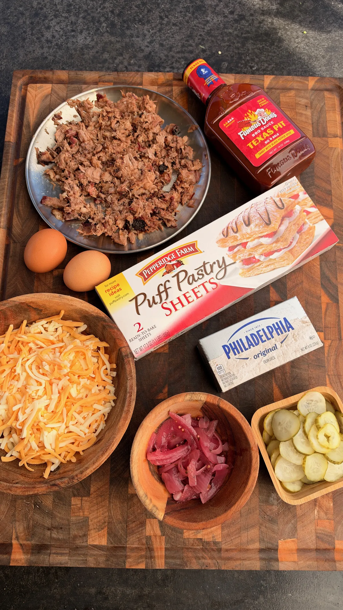 Flat lay of smoked brisket hot pocket ingredients: puff pastry, cream cheese, shredded cheddar, pickles, pickled red onions, BBQ sauce, and eggs