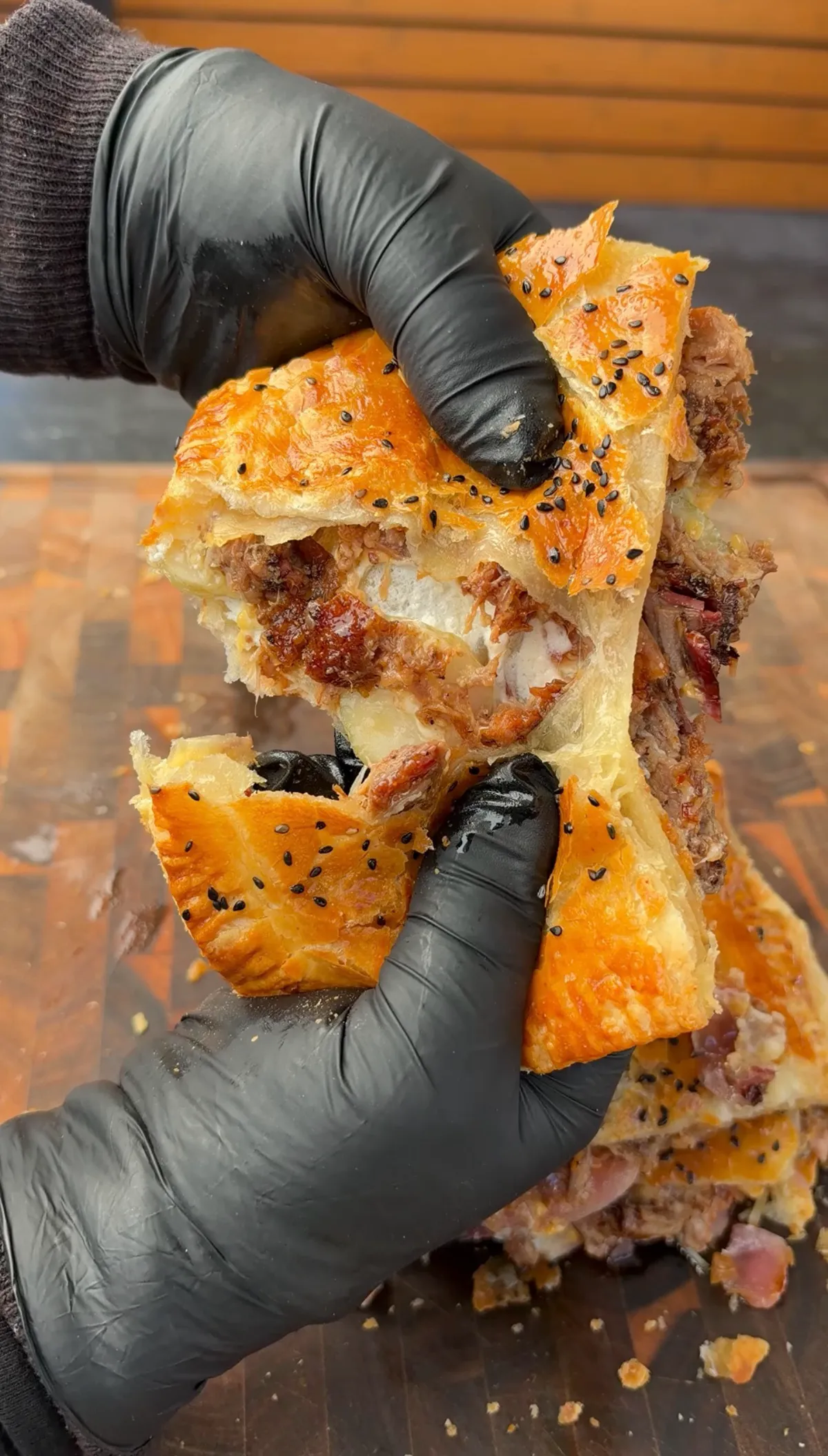 Hands in black gloves pulling apart golden puff pastry hot pocket filled with smoked brisket, cheddar, pickles, and BBQ sauce