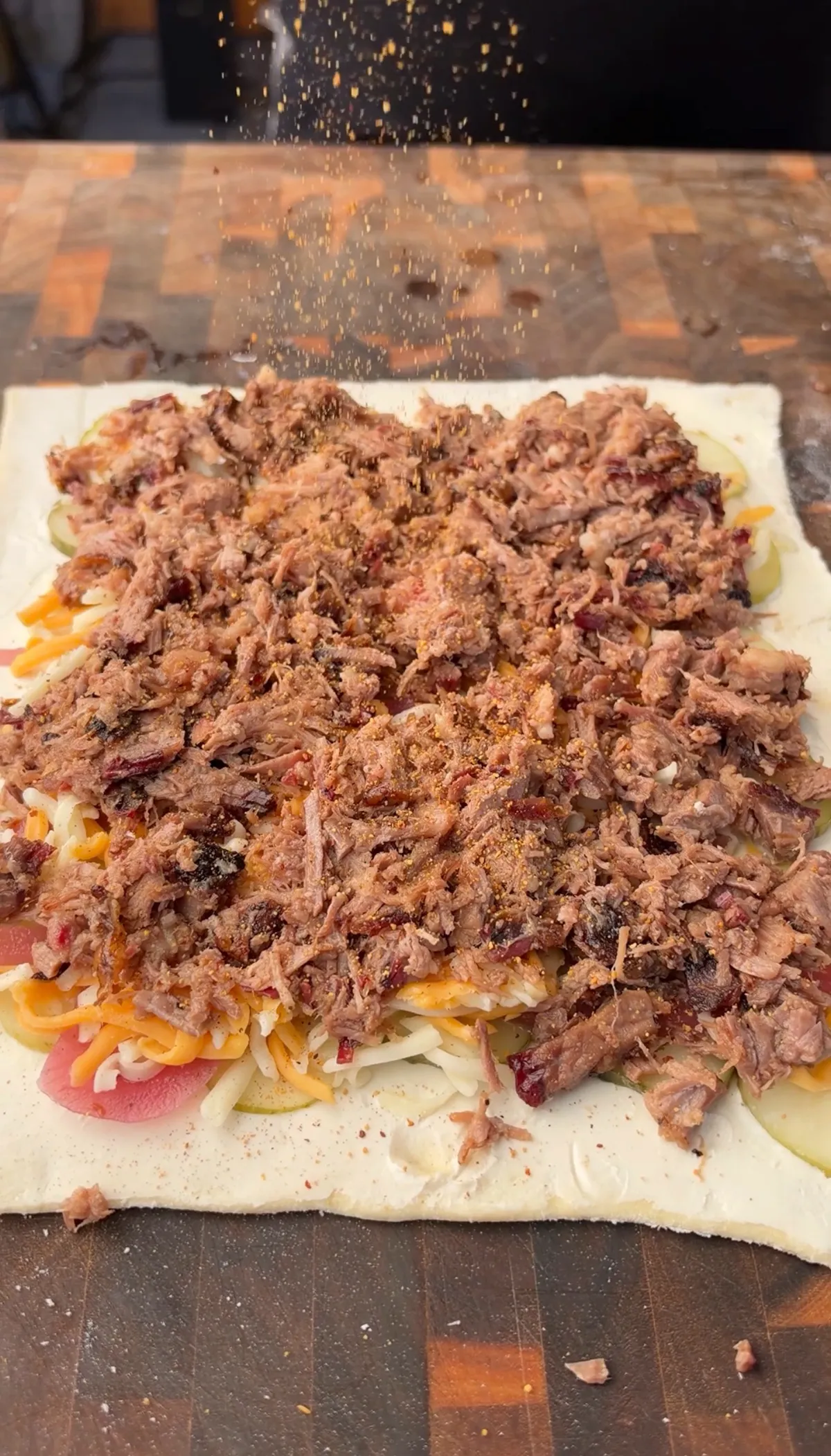 Sweet BBQ rub being sprinkled over chopped smoked brisket layered on puff pastry with cheddar, pickles, and pickled red onions