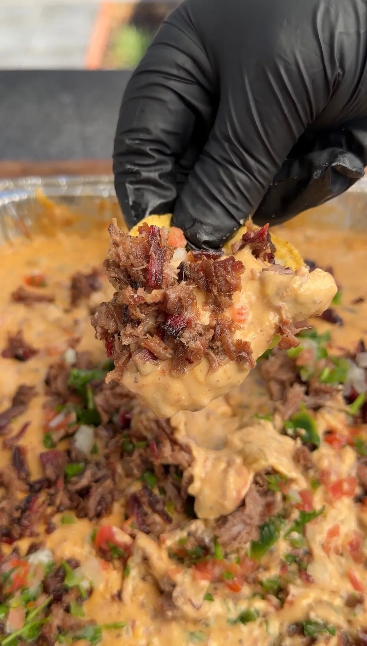 Gloved hand holding a tortilla chip loaded with smoked brisket queso topped with chopped brisket and pico de gallo