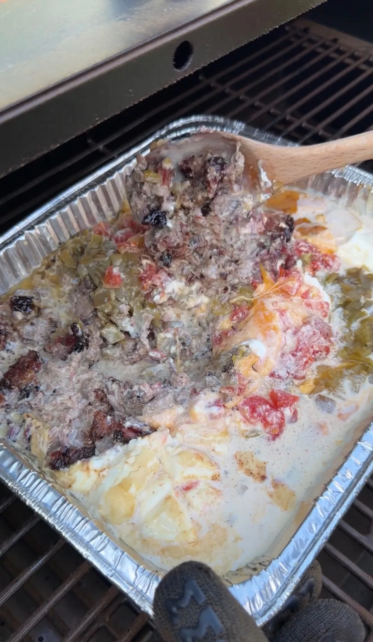Smoked queso with chopped brisket, Rotel, green chiles, and melted Velveeta being stirred in a foil tray on a smoker