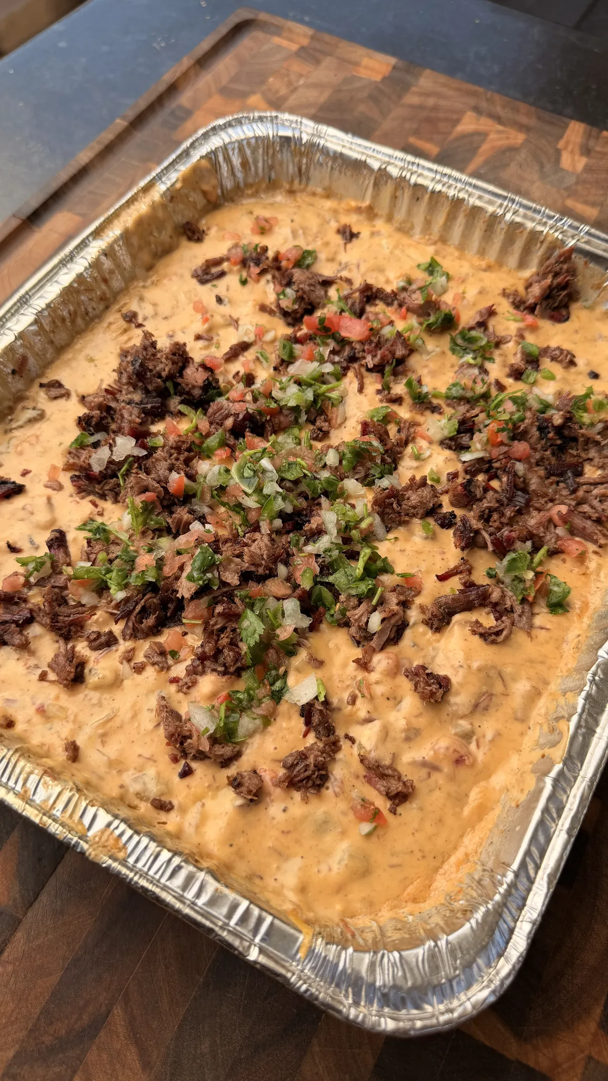 Smoked queso in foil tray topped with chopped brisket, fresh pico de gallo, and cilantro on a wood board