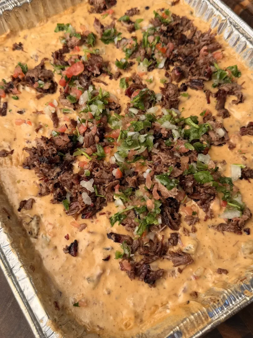 Smoked queso in foil tray topped with chopped brisket, fresh pico de gallo, and cilantro