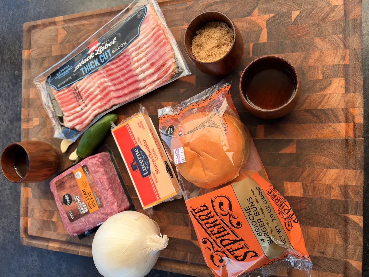 Ingredients for smoked cheddar stuffed burgers with bacon jam: ground beef, thick cut bacon, cheddar cheese, brioche buns, yellow onion, jalapeño, brown sugar, and maple syrup on a wood cutting board