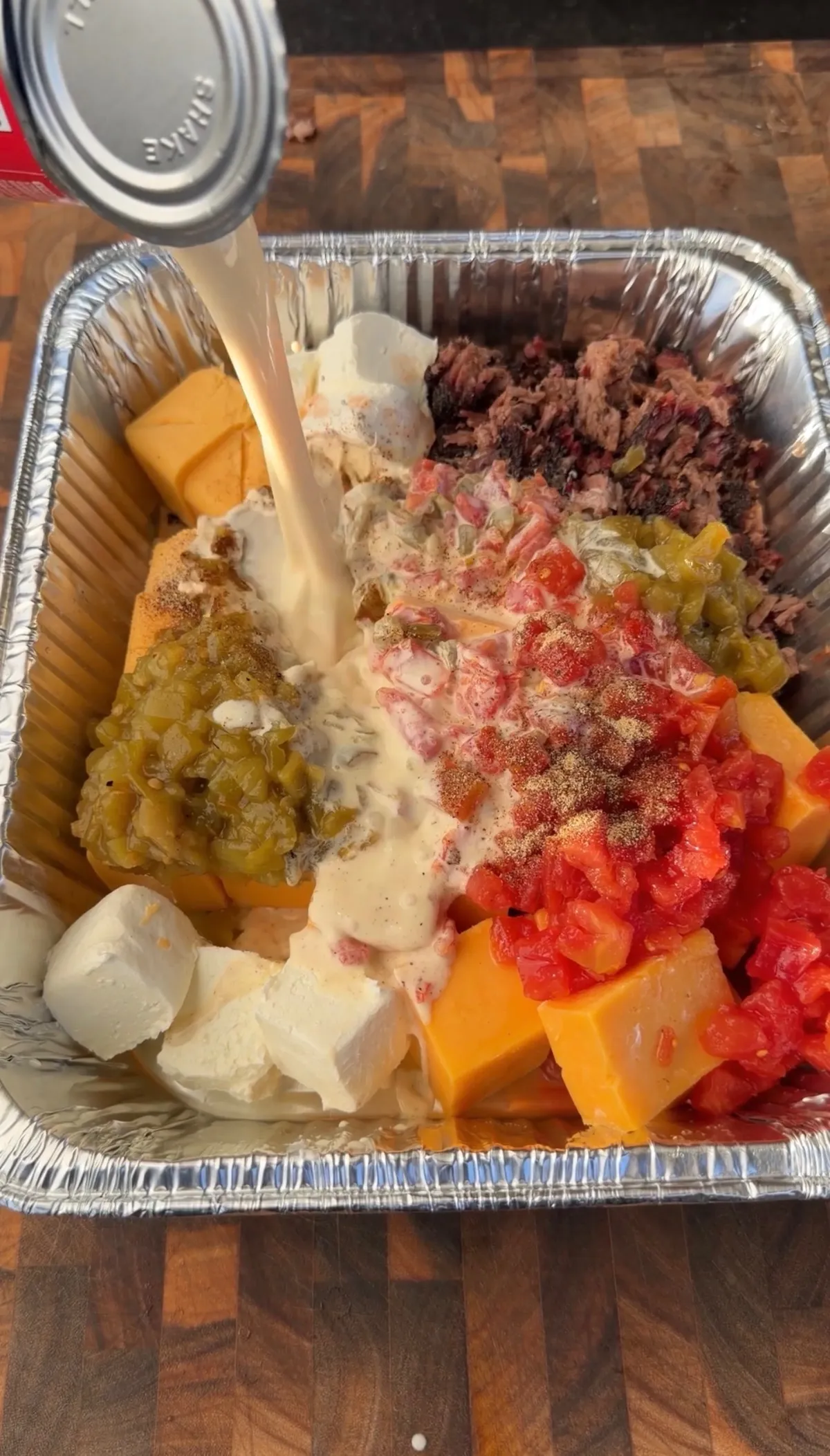 Milk being poured into foil tray with Velveeta, cheddar, Rotel, green chiles, and chopped brisket for smoked queso
