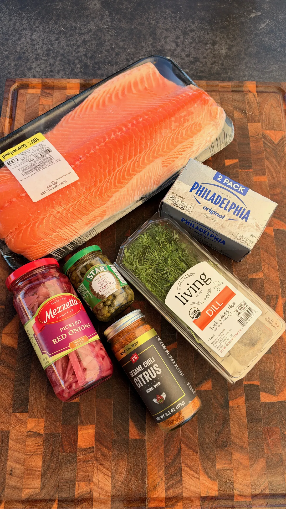 Smoked salmon dip ingredients: salmon fillet, Philadelphia cream cheese, capers, pickled red onions, fresh dill, and sesame chili citrus seasoning on a wood cutting board