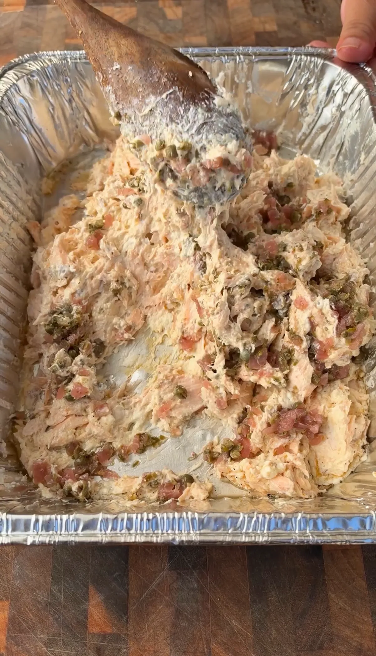 Smoked salmon and cream cheese dip being mixed with capers and pickled red onion in a foil pan