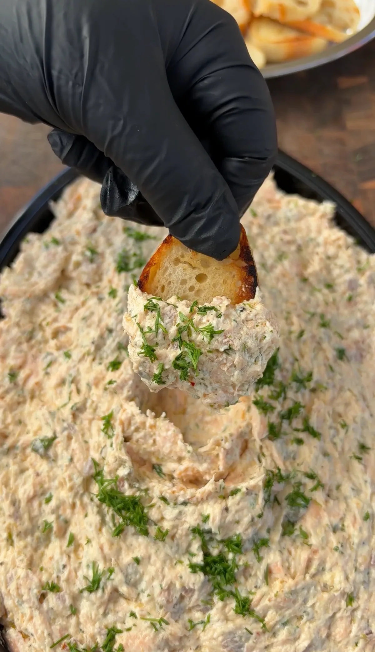 Gloved hand dipping toasted bread into smoked salmon cream cheese dip garnished with fresh dill in a black skillet