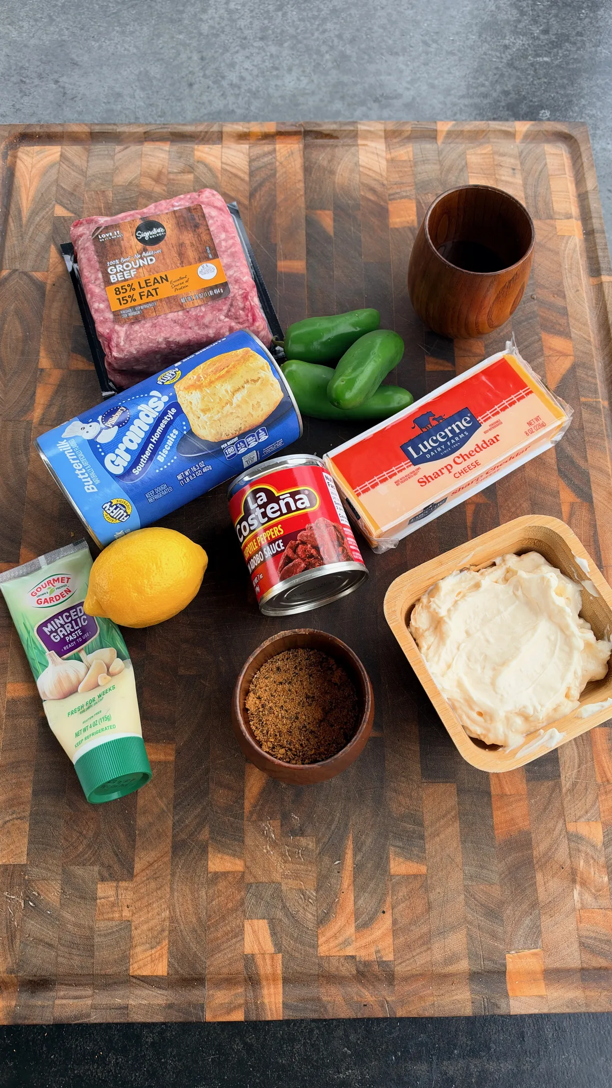 Ingredients for Smoked Spicy Cheeseburger Bombs: ground beef, Grands biscuit dough, jalapeños, cheddar cheese, chipotle sauce, garlic paste, mayo, lemon, and seasoning on a wood cutting board