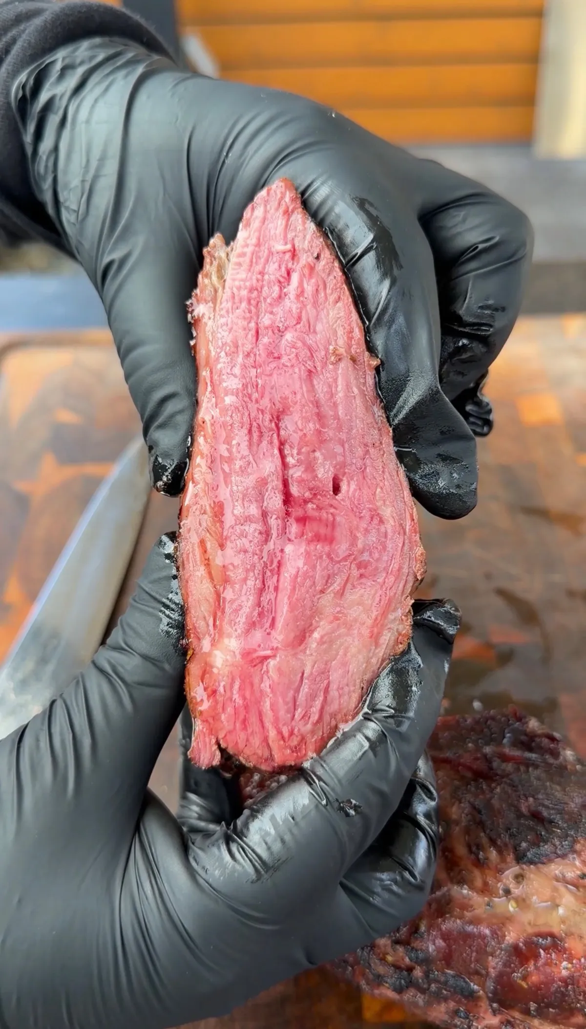 Hands in black gloves holding a slice of reverse-seared smoked tri-tip showing a pink medium-rare interior with smoky bark