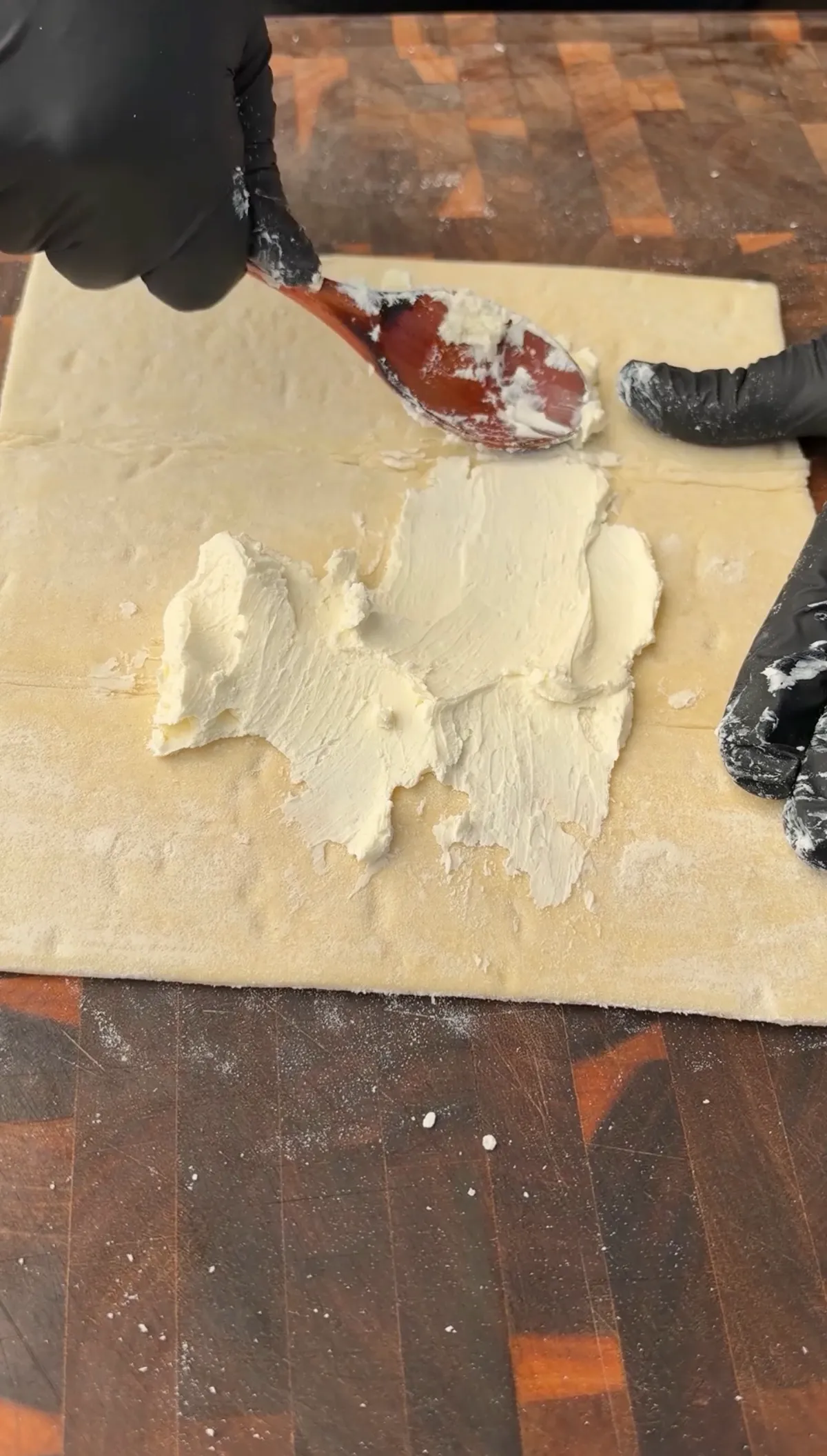 Gloved hands spreading softened cream cheese evenly over a sheet of thawed puff pastry on a floured wood cutting board