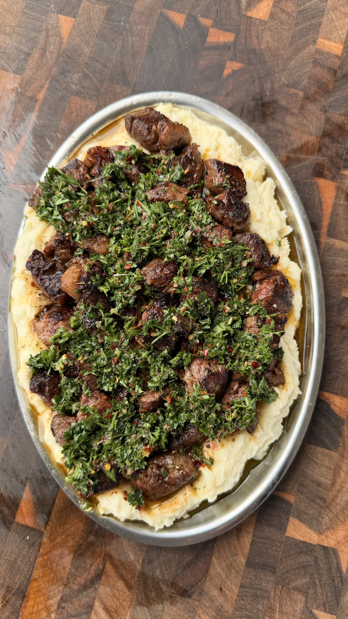 Pan-seared ribeye steak bites topped with fresh parsley chimichurri served over creamy mashed potatoes on a silver oval platter