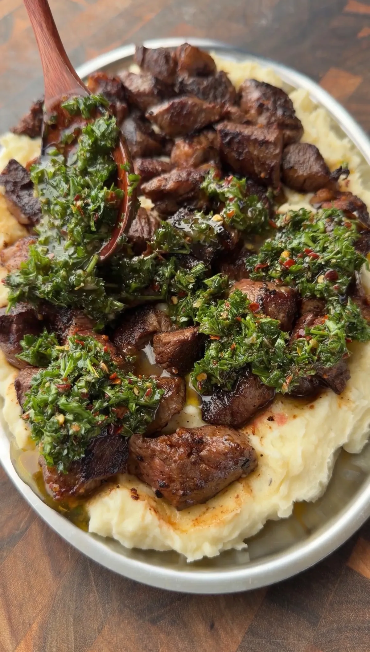 Pan-seared ribeye steak bites over creamy mashed potatoes topped with fresh parsley chimichurri in a white bowl