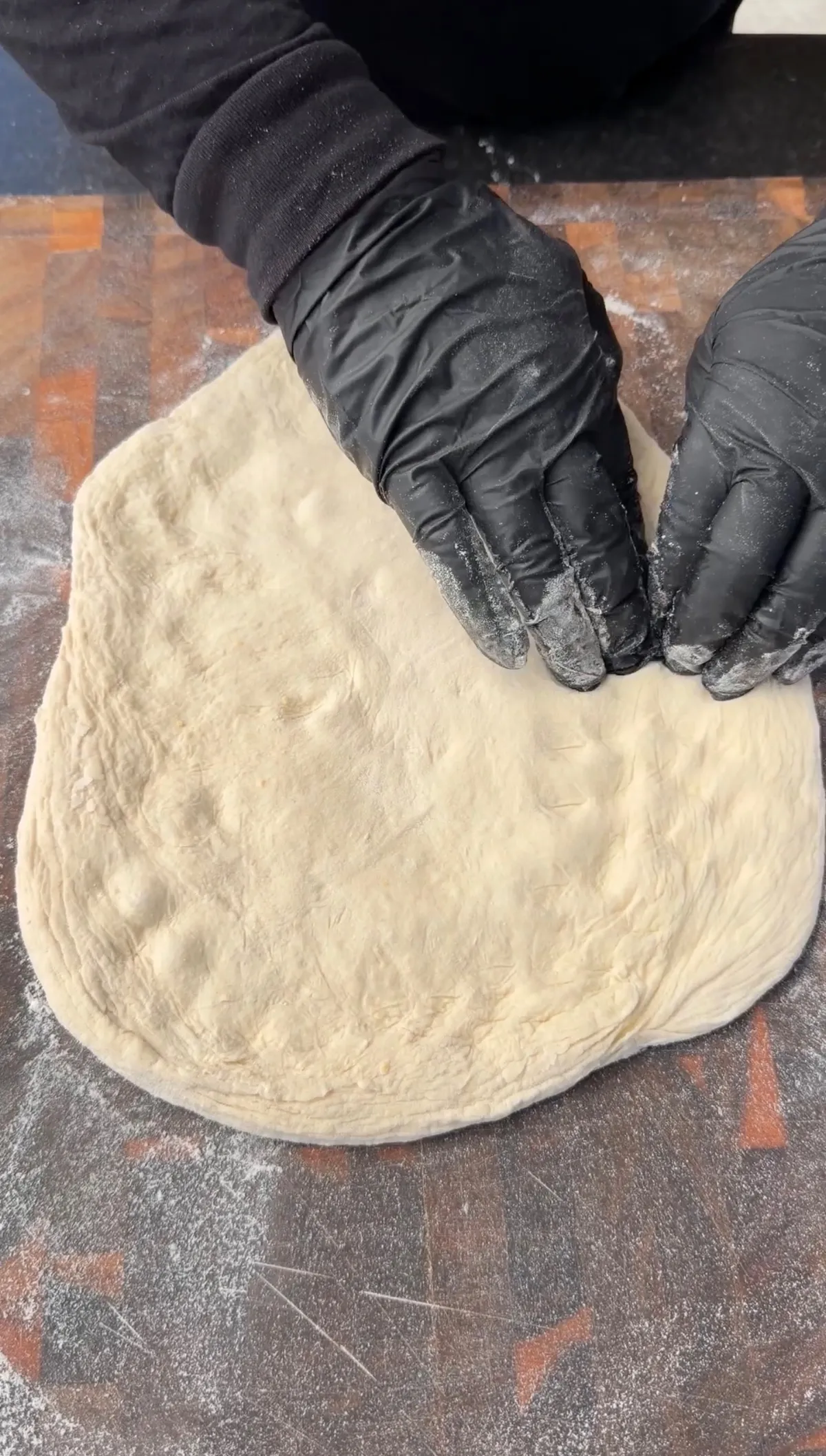 Hands in black gloves stretching pizza dough on a floured surface to prepare a pizza sandwich