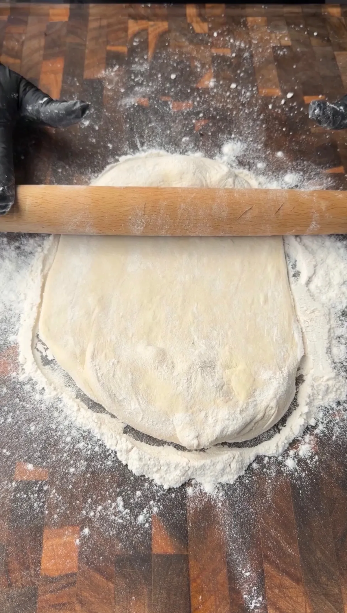 Pizza dough being rolled into a round shape with a wooden rolling pin on a floured wooden surface for a calzone
