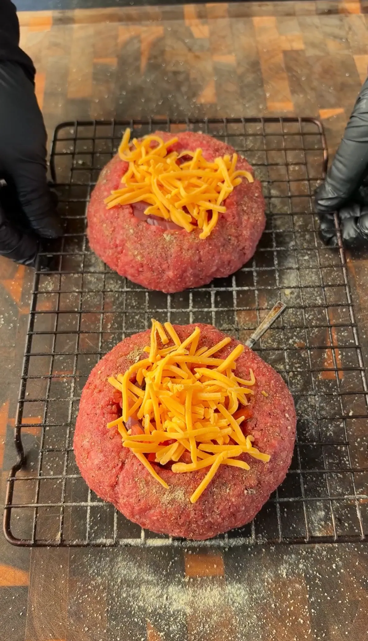 Two raw 85/15 ground beef patties with Mason jar indentations filled with freshly grated cheddar cheese on a wire rack