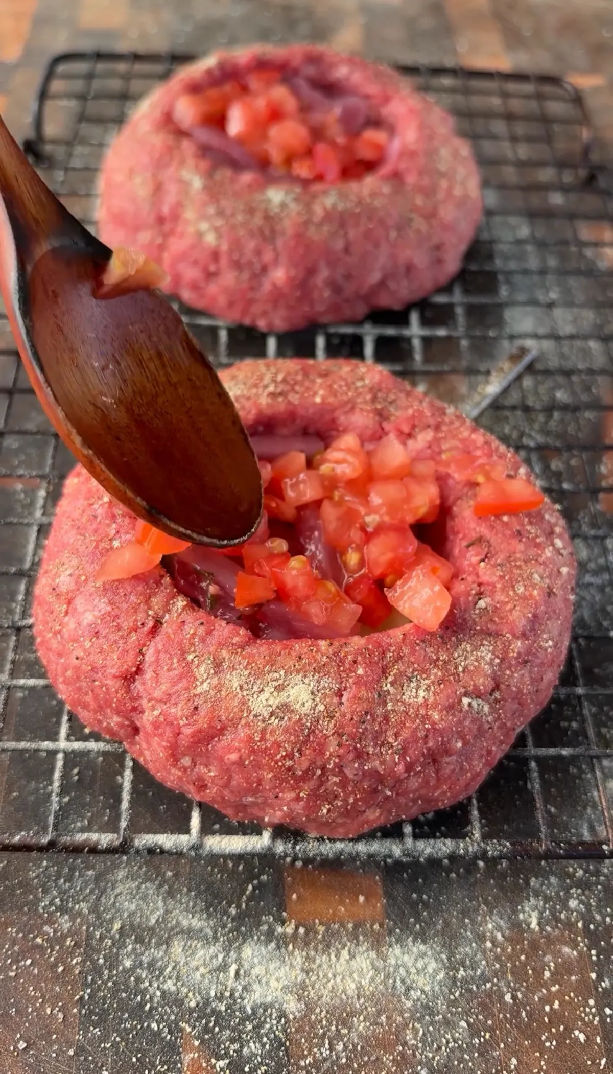Stuffing raw beer can burger patties with diced tomatoes and pickled red onions using a wooden spoon on a wire rack