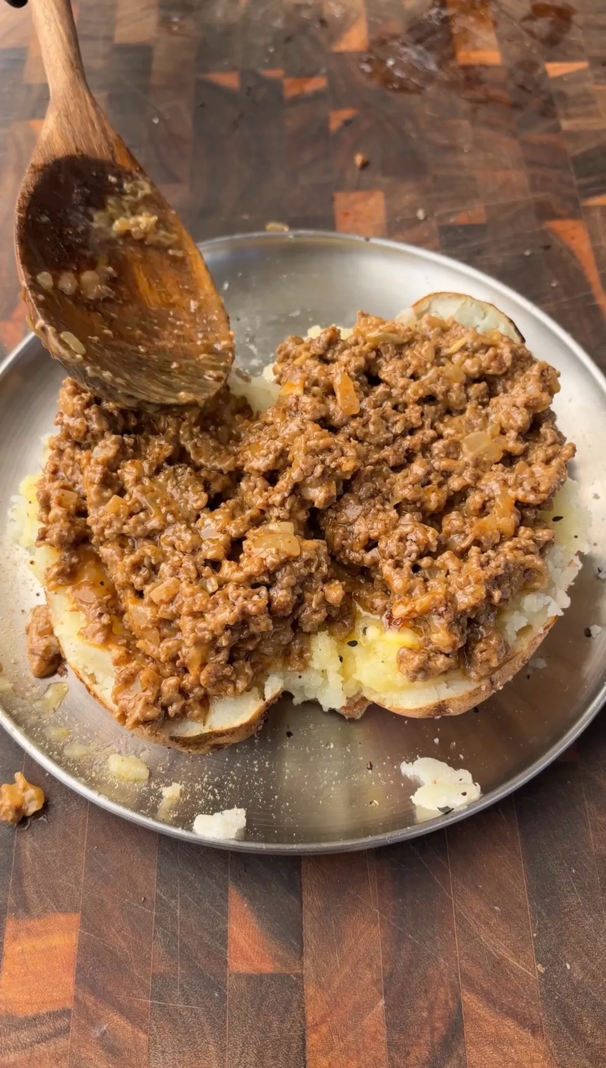 Taco beef with nacho cheese and hot sauce being spooned over a fluffed russet potato half on a silver plate