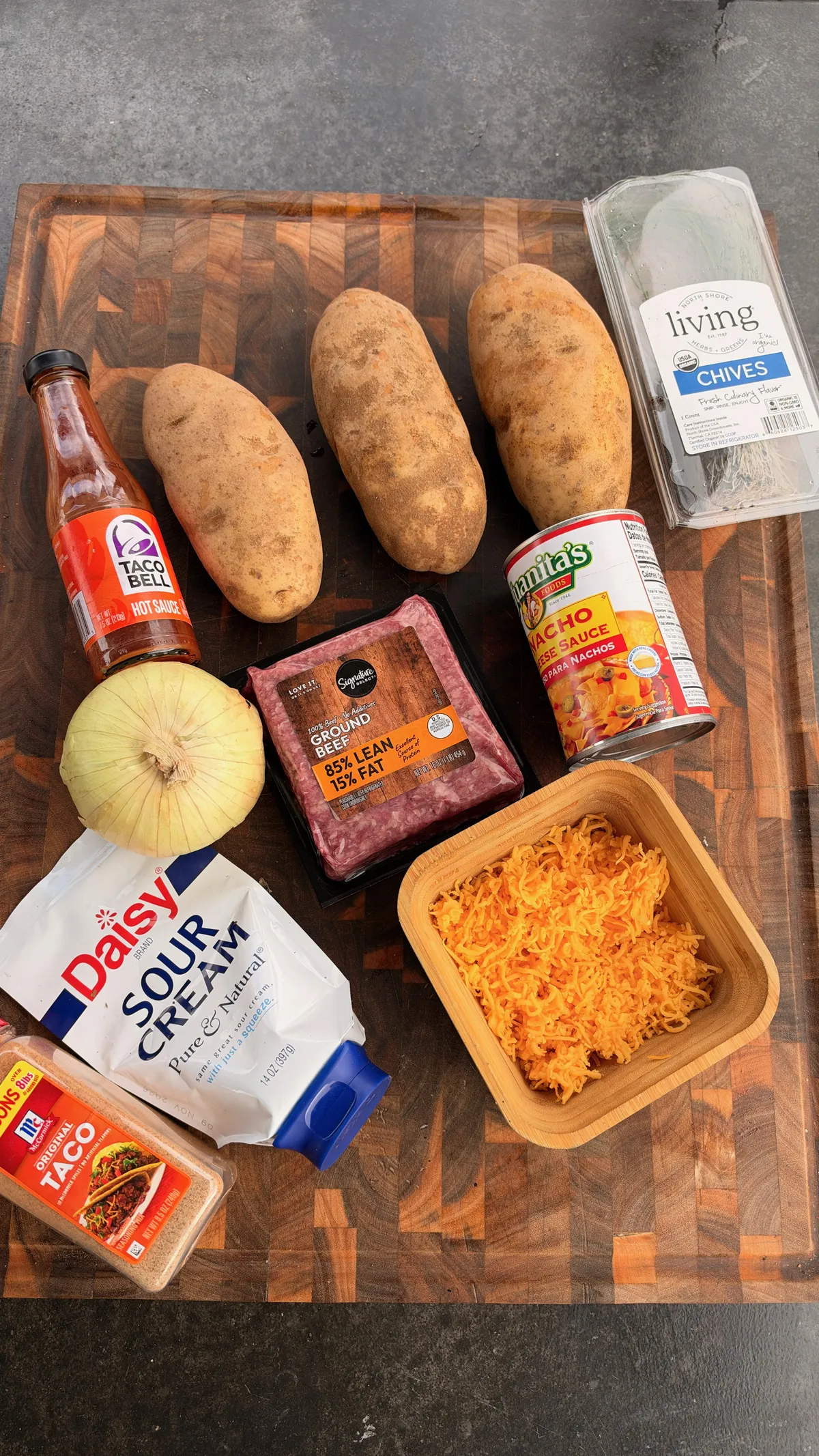 Flat lay of taco loaded baked potato ingredients: russet potatoes, 85/15 ground beef, nacho cheese, sour cream, cheddar, chives, taco seasoning, and Taco Bell hot sauce